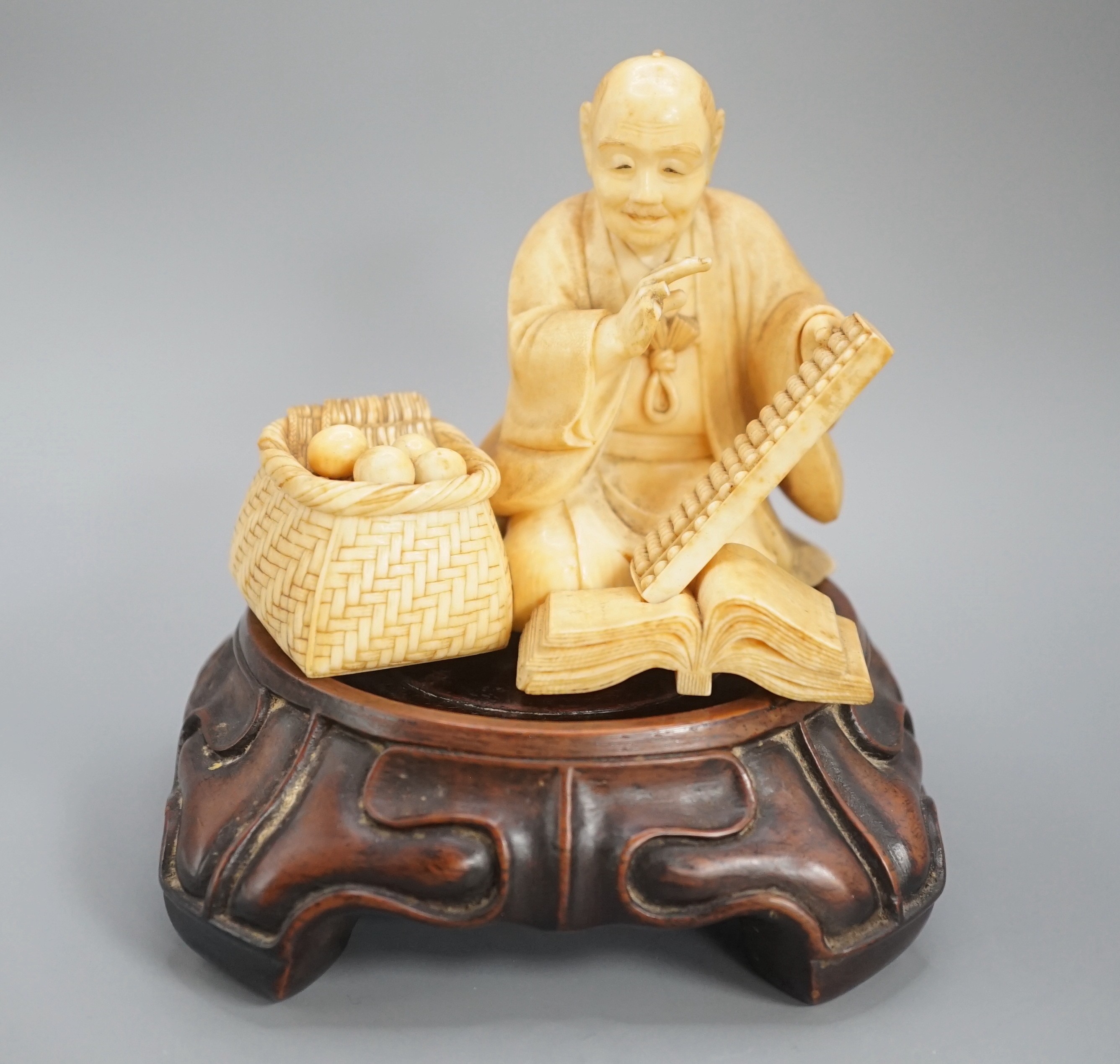 A Japanese ivory okimono of a street vendor kneeling and holding an abacus, Meiji period, 8cm, associated wood stand